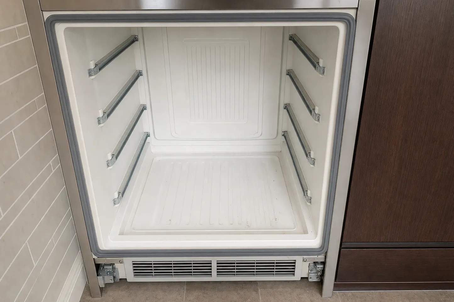 Emptied undercounter refrigeration cavity: cream liner, shelf rails on both walls, door gasket and a floor-level vent grille