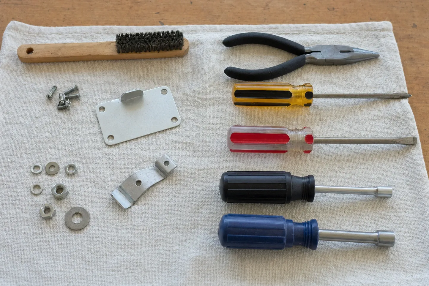 Overhead view of four drivers, needle-nose pliers and a wire brush on white cloth with a sheet-metal cover plate and washers